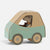 A small Maileg mouse dressed as a driver inside a handmade cardboard car. The car features a sage green side panel, a natural brown cardboard roof and wheels, and a small embroidered patch on the side.