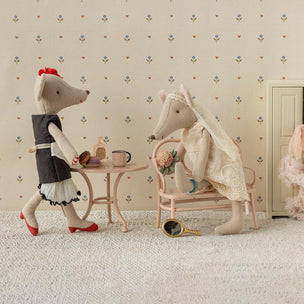Two Maileg mice in a room with a small table and chair, set against a patterned wallpaper.