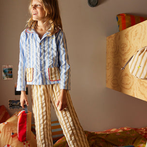 Person wearing a patterned outfit standing in a bedroom with colorful pillows and furniture.