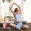 Woman practicing yoga outdoors on a wooden deck with a pink water bottle nearby.
