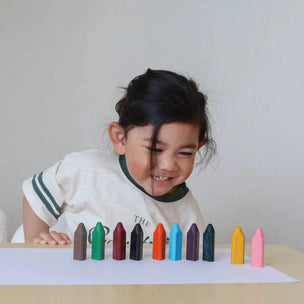 A young child with dark hair smiles while looking at a row of colorful crayons lined up on a white sheet of paper on a light wooden table.