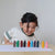 A young child with dark hair smiles while looking at a row of colorful crayons lined up on a white sheet of paper on a light wooden table.