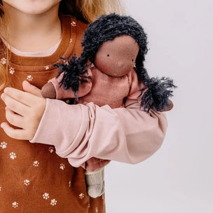 A child in a brown patterned top holds a soft fabric doll with dark curly hair and a dusty pink body against a plain light gray background.