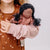 A child in a brown patterned top holds a soft fabric doll with dark curly hair and a dusty pink body against a plain light gray background.