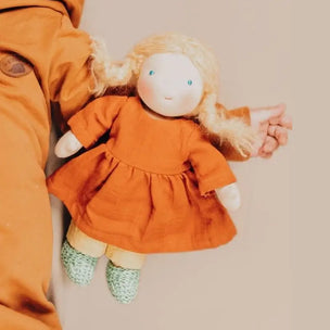 A handmade fabric doll with curly blonde hair, wearing an orange dress, cream pants, and green patterned shoes, held by a person wearing an orange jacket against a plain beige background.