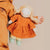 A handmade fabric doll with curly blonde hair, wearing an orange dress, cream pants, and green patterned shoes, held by a person wearing an orange jacket against a plain beige background.