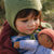 Child wearing a green knitted bonnet holding a brown doll outdoors.