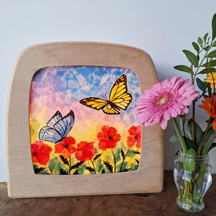 Decorative piece with butterflies and flowers in a wooden frame, next to a vase with flowers.