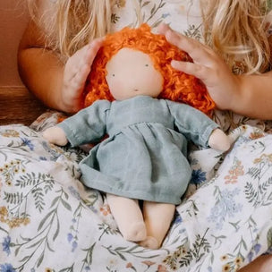 A handmade rag doll with curly orange hair and a dusty blue dress, held gently by hands over a floral blanket.