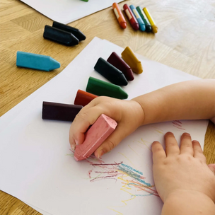 A child's hands drawing with pink and blue crayons on white paper over a light wood table, with other crayons scattered nearby.