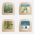 Four square wooden-framed art pieces arranged in a 2x2 grid on a white background. Each piece features a different painted scene: a field scene with children, a beach walk with children, an autumn tree scene, and a snowy winter scene.