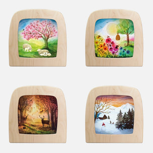 Four square wooden picture frames arranged in a 2x2 grid on a white background, each containing a different colorful landscape painting: a pink tree with sheep, flowers and a bee, a forest with deer, and a snowy winter scene.