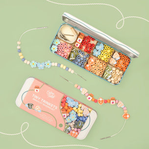 A flat-lay product shot of a 'Tiny Trinkets Needle Trinket Set' by The Happy Tinker. The set includes a silver metal tin filled with colorful seed beads, a pink and white booklet, and a decorative bead strand with needles, all arranged on a light sage green background.