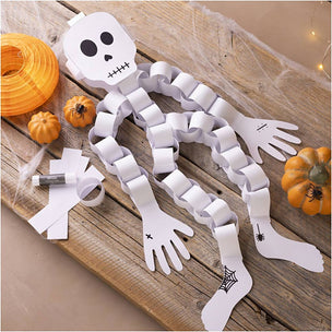 White paper skeleton Halloween decoration laid out on a rustic wooden surface with orange pumpkins, fake spiderwebs, and small orange lanterns nearby.