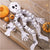 White paper skeleton Halloween decoration laid out on a rustic wooden surface with orange pumpkins, fake spiderwebs, and small orange lanterns nearby.