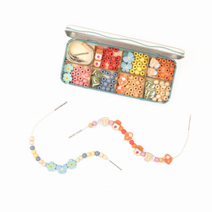 A bead-making kit shown open with a variety of colorful seed beads in different shapes and sizes, along with two beaded cords and metal needles, on a plain white background.