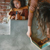 Grapat Advent Calendar 2025 Two children playing with a toy train set on a concrete surface against a stone wall.