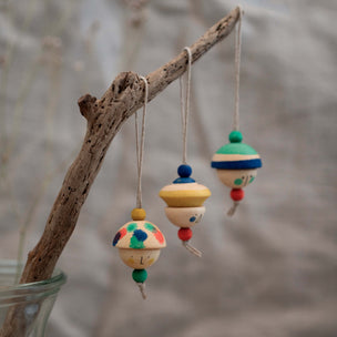 Three hand-painted wooden kind folk faces hang from a natural branch with string, set against a soft, blurred neutral background.