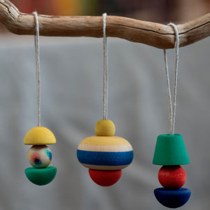 Three colorful wooden hanging ornaments with painted geometric shapes suspended from a natural wood branch against a soft blurred gray background.