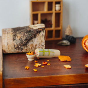 A styled tabletop scene with a rolled parchment letter, small orange and gold artifacts, a birch log, and a partially peeled orange on a wooden surface against a soft gray background.