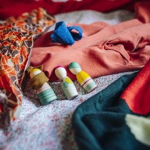 Three small hand-painted wooden peg dolls with colorful painted heads and bodies rest on a bed surrounded by various colorful fabrics, including a rust-colored shirt, a red and black patterned textile, and a dark teal knit blanket.