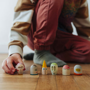 A child wearing a colorful knit sweater and rust-colored pants sits on a wooden floor, reaching toward a row of small hand-painted wooden toy figures.