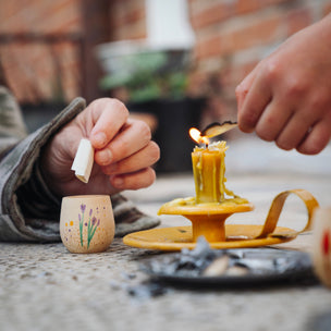A close-up lifestyle shot of a person lighting a yellow candle in a brass holder outdoors. Another hand holds a small white stick, and a small hand-painted cup with purple flowers sits nearby on a stone surface.