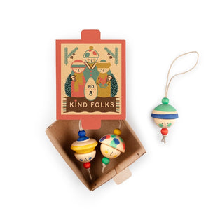 A wooden dreidel toy set shown with its illustrated cardboard box on a white background. The open box contains two small wooden dreidels, and a third dreidel hangs to the right by a string.