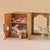 Miniature dollhouse with open wooden wardrobe, plush toys, and small table with chair on a beige background.