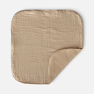 A square muslin baby blanket in a warm beige color with a subtle woven grid pattern, shown on a light gray background with one corner folded over to reveal the soft fleece backing.