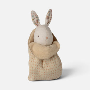 A plush stuffed bunny wrapped in a soft hooded blanket, photographed on a clean light gray studio background. The bunny has pale cream fabric, patterned ears with small floral motifs, and a beige fleece hood.