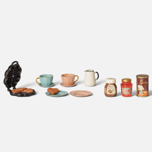 A miniature Maileg toy breakfast set arranged on a light gray background, including a black waffle iron with cookies, a light blue cup, a peach cup and saucer, a white pitcher, two small plates, two jars, and a can of coffee.