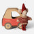 A Maileg brand toy set featuring a small mouse doll wearing a red and beige striped sweater and red hat, standing next to a red and brown cardboard car with the Maileg logo.