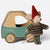 A Maileg brand plush mouse toy dressed as a gnome, standing next to a wooden toy car. The mouse has a red felt hat, a brown and beige striped knit sweater, and green pants, holding a light pink string.