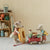 A styled indoor scene with two plush mouse figures dressed in picnic clothing, one carrying a striped donut and the other pushing a red wagon loaded with picnic items, beside a wooden shelf with camping accessories and a lantern.