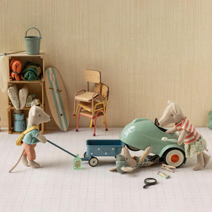 A styled miniature scene with small animal figurines and toy props arranged on a white floor against a beige wall. One figurine pushes a blue wagon while another stands beside a mint green toy car, with additional beach-themed accessories and furniture in the background.