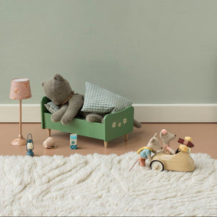 A styled children's bedroom scene with a green wooden bed frame holding a gray stuffed bear and pillows, a pink lamp, a small green cabinet, and a wooden mouse toy driving a car on a white shag rug against a pale green wall.