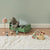 A styled children's bedroom scene with a green wooden bed frame holding a gray stuffed bear and pillows, a pink lamp, a small green cabinet, and a wooden mouse toy driving a car on a white shag rug against a pale green wall.