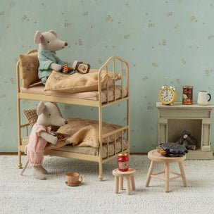 A miniature dollhouse-style room scene with two plush mouse figures arranged on a wooden bunk bed, alongside small furniture pieces, a wallpapered wall, and a fireplace display.
