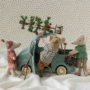 Three plush Maileg toy mice dressed in winter clothing gather around a small teal toy truck on white fabric, with one mouse loading a decorated pine branch into the truck bed. The background is a cream fabric with small blue polka dots.
