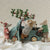 Three plush Maileg toy mice dressed in winter clothing gather around a small teal toy truck on white fabric, with one mouse loading a decorated pine branch into the truck bed. The background is a cream fabric with small blue polka dots.
