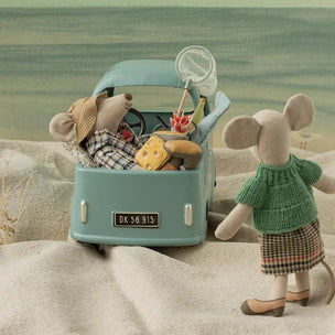 A styled miniature scene featuring a small plush mouse standing beside a light blue toy car filled with beach-themed toys and snacks, set on beige sand against a soft blue-green background.