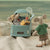A styled miniature scene featuring a small plush mouse standing beside a light blue toy car filled with beach-themed toys and snacks, set on beige sand against a soft blue-green background.