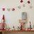 A styled tabletop display against a plain wall with a knitted heart and bell garland hanging above. The scene includes a small doll on the left, several animal figurines, candles, wrapped gift boxes, and a tiny Christmas tree, creating a cozy decorative setup.
