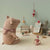 A styled collection of plush toys and miniature accessories arranged on a light wood floor against a pale green wall. The set includes a large pinkish-beige pig plush wearing a red-and-white gingham scarf, a gray mouse plush, a small red-and-white dressed doll, a miniature lamp, chair, and framed picture, plus tiny scattered gold coins.