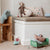 A styled children's room corner featuring a white built-in bench with a patterned cushion, a plush monkey doll, and pillows. To the left is a wooden crate shelf holding a blue plush elephant, and in the foreground is a small green toy bed with stuffed cushions on a light wood floor.