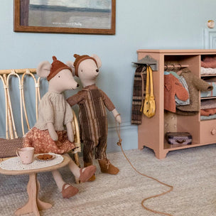 A styled interior scene with two Maileg plush mouse dolls posed beside a small wooden bench and table, next to a pastel pink clothing rack filled with folded garments and accessories. The room has pale blue walls, a textured rug, and a framed painting above the bench.
