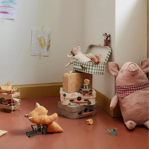 A styled children's room corner with a pink plush pig chair, stacked vintage-style suitcases, a stuffed animal on a green-and-white checkered cushion, pillows, and small toys on a terracotta floor against cream walls.