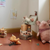 A styled children's room corner with a pink plush pig chair, stacked vintage-style suitcases, a stuffed animal on a green-and-white checkered cushion, pillows, and small toys on a terracotta floor against cream walls.
