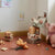 A styled children's room corner with a pink plush pig chair, stacked vintage-style suitcases, a stuffed animal on a green-and-white checkered cushion, pillows, and small toys on a terracotta floor against cream walls.